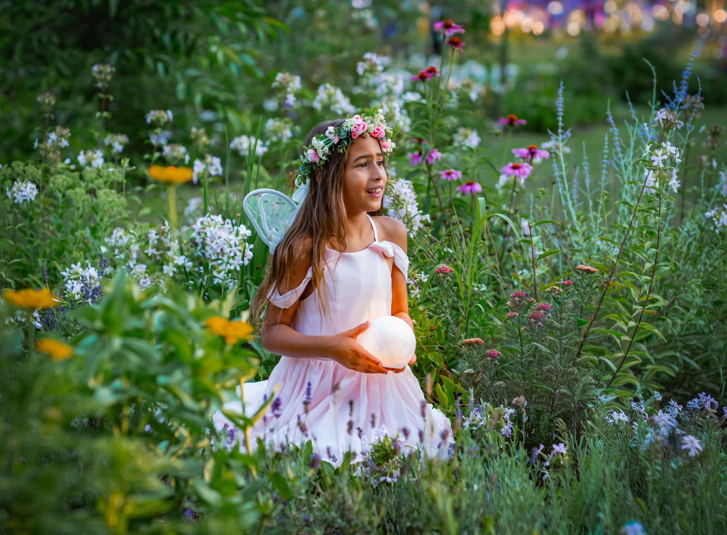 Young girl dressed as a fairy with wings and flower crown holds glowing orb in a lush garden.
