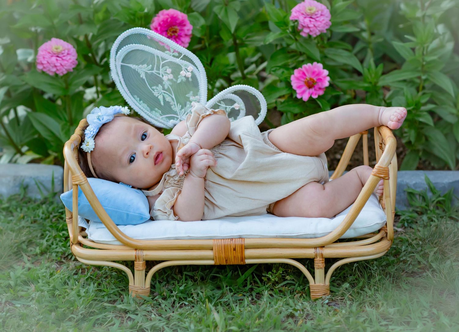 Baby in cream onesie with fairy wings lying on rattan bed outdoors surrounded by pink zinnia flowers.