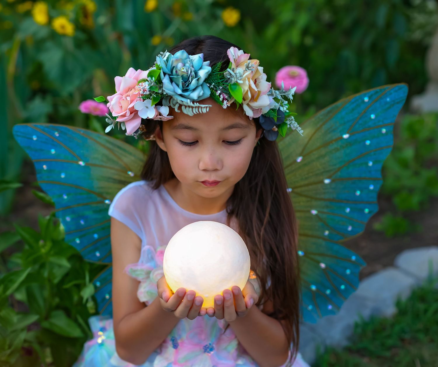 A young girl dressed as a fairy with blue wings and floral crown holds a glowing orb in a garden.