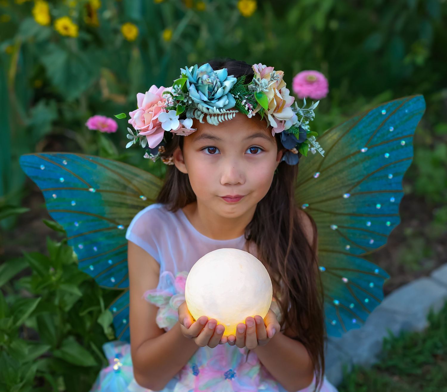 A young girl dressed as a fairy with blue wings and floral crown holds a glowing orb in a garden.