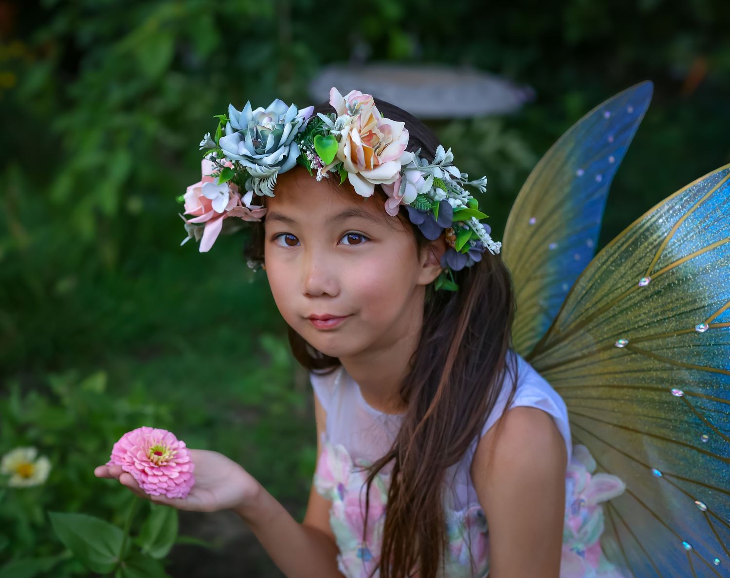 Young girl dressed as a fairy with butterfly wings and floral crown holds a pink zinnia flower in a garden.