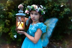 A young girl dressed as a fairy in a blue dress, flower crown, and wings holds a glowing lantern outdoors.