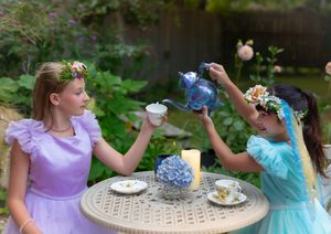 Two girls in princess dresses enjoy a garden tea party, one pouring from a blue teapot outdoors.