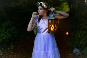 Girl in white tulle dress and flower crown holds glowing lantern in enchanted forest path at dusk.