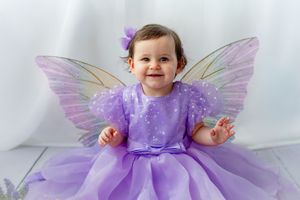 Smiling baby girl in purple fairy costume with butterfly wings and bow, sitting against white background.