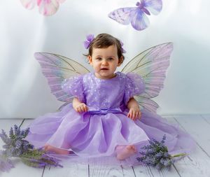 Baby girl in purple fairy costume with iridescent wings sits among lavender flowers with butterflies in background.
