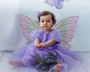Baby girl in purple fairy dress with wings holds lavender bouquet, sitting on white floor.