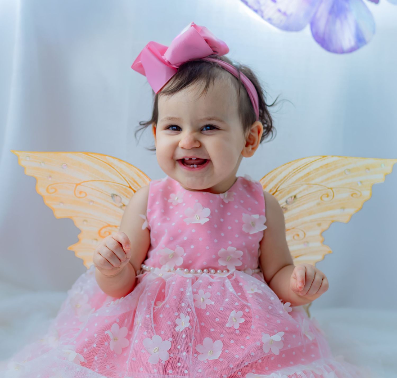 Smiling baby girl in pink dress with butterfly wings and pink bow headband poses for a portrait photo.