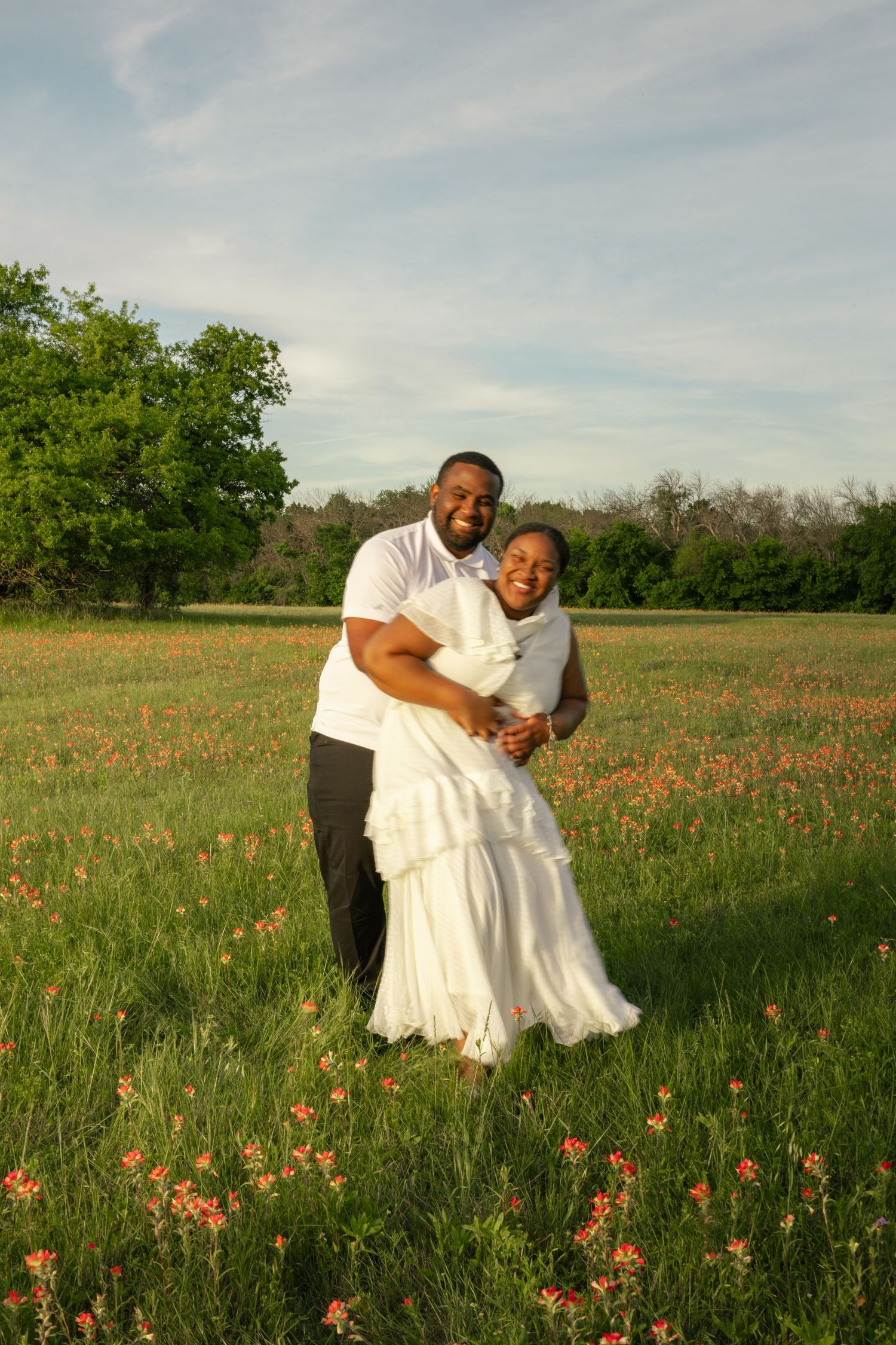 Happy couple embracing in a wildflower meadow, woman in white dress, man in white shirt, green trees and blue sky behind them.