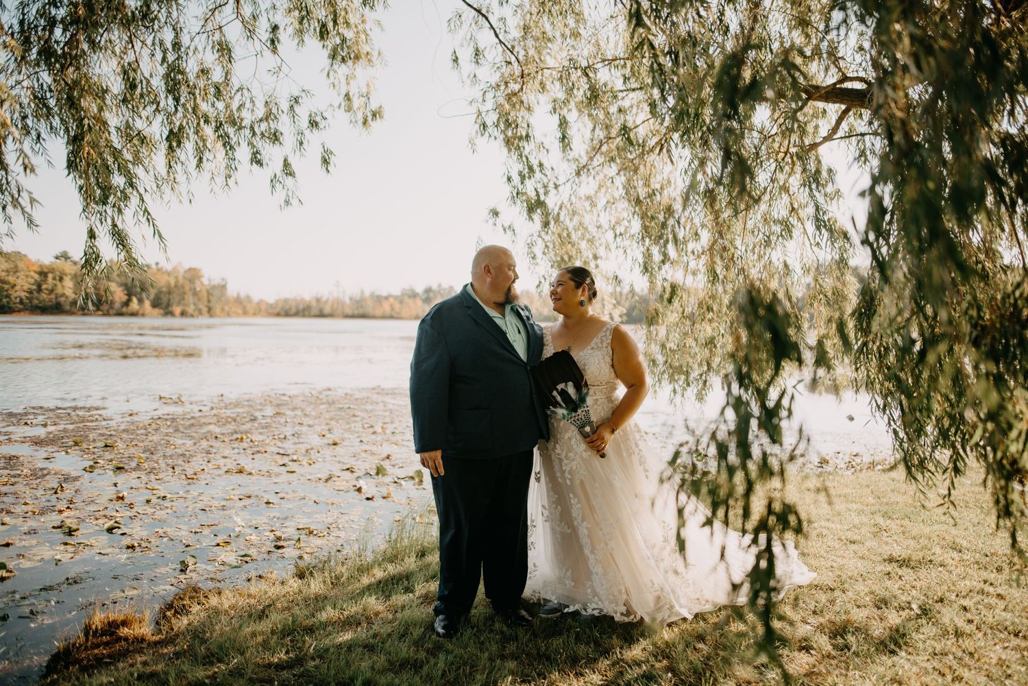 A romantic autumn wedding photo under willow branches by a serene lake with fallen leaves scattered on the ground.