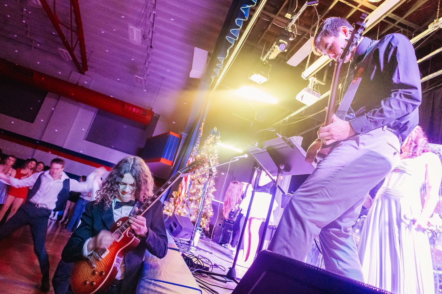Live band performing energetically on stage at an indoor venue with colorful lights and dancing guests in the background.