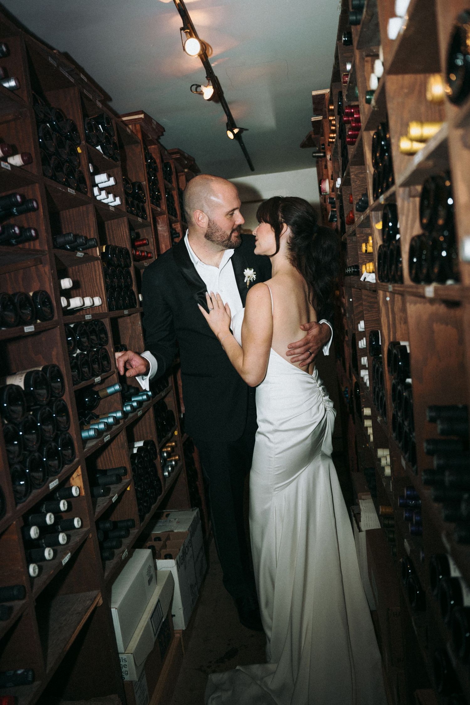 Bride and groom share a romantic moment in a wine cellar, surrounded by wooden racks filled with bottles.