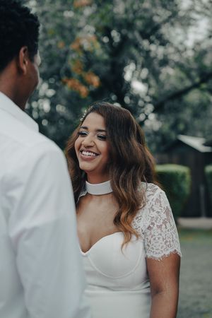 Radiant bride in lace wedding dress smiles joyfully at her groom in an outdoor garden setting.