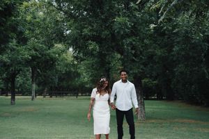 Interracial newlywed couple walking hand in hand through a lush green park on their wedding day.