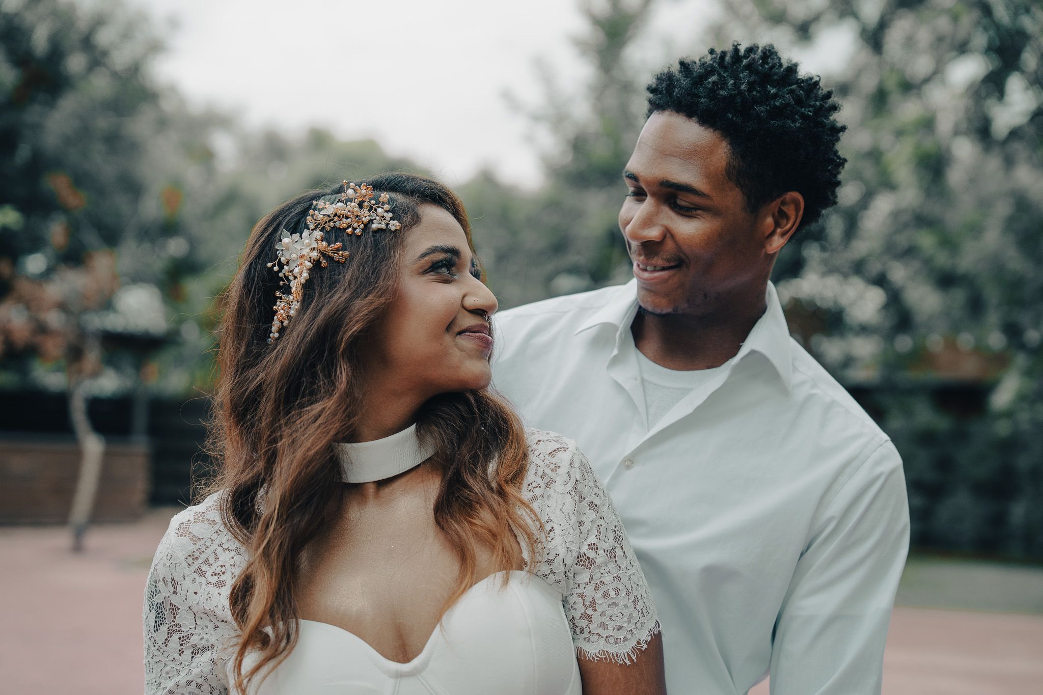 Interracial couple smiling at each other outdoors, bride in white lace dress wearing a choker and gold floral headpiece.