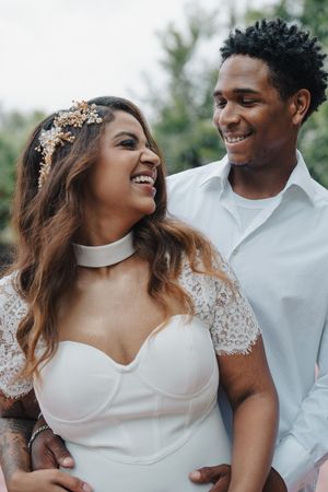 Laughing bride in white lace sweetheart dress with gold floral headpiece embraced by smiling groom outdoors.