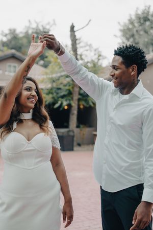 Joyful interracial couple dancing together outdoors at their wedding, groom spinning bride in white dress.
