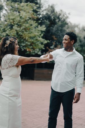 Joyful interracial wedding couple holding hands and dancing outdoors on a brick path surrounded by green trees.