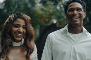 Happy interracial couple smiling together outdoors, bride wearing white lace dress with floral hair accessory.