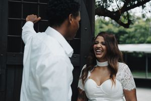 Bride in white lace dress laughing with groom in white shirt near a dark wooden door during wedding photoshoot.