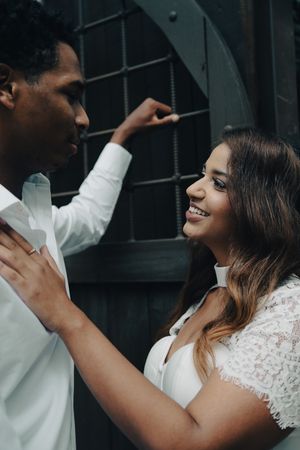 Bride in white lace dress smiling up at groom while touching his chest near a dark ornate door backdrop.