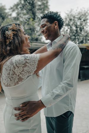 Interracial wedding couple embracing and smiling at each other outdoors, bride in lace dress with gold floral headpiece.