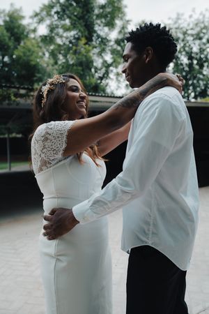 Bride in white lace wedding dress with gold crown smiling at groom while embracing outdoors in a park.
