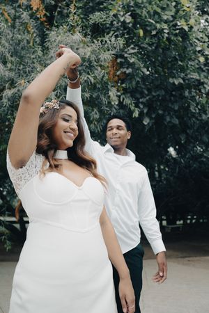 Bride in white lace dress with floral crown dancing with groom outdoors, arm raised joyfully in celebration.