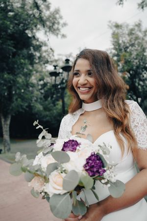 Smiling bride in white lace bolero gown holding lush bouquet of white roses and purple flowers outdoors.