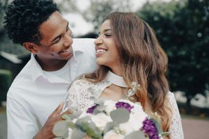 Smiling interracial couple in wedding attire sharing a tender moment outdoors, bride holding a bouquet of white and purple flowers.