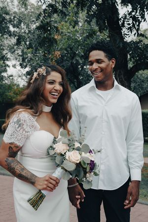 Laughing bride in white lace dress with floral hair accessory and groom in white shirt holding bouquet in a park setting.