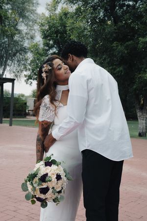 Groom in white shirt kissing tattooed bride on cheek, bride holding white and purple floral bouquet outdoors.