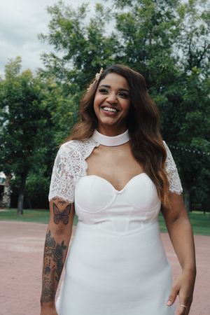 Smiling tattooed bride in white sweetheart gown with lace bolero and choker necklace posing outdoors.