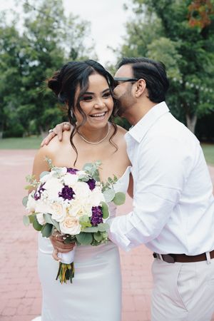 Laughing bride holding white and purple bouquet as groom whispers in her ear in a green park setting.