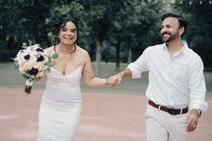 Bride and groom walking hand in hand outdoors, bride in white gown holding purple and white floral bouquet.