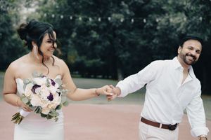 Bride holding bouquet laughs while groom leads her by hand in park.