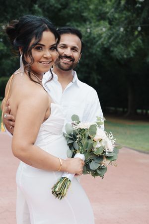 Smiling groom embraces bride holding white and green bouquet in park.