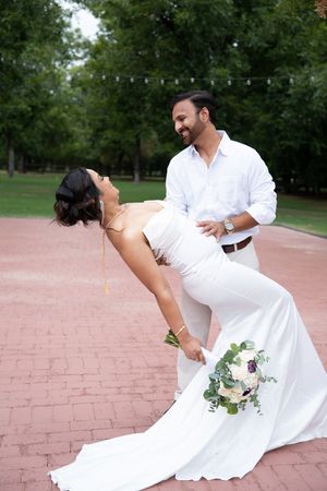 Groom dipping bride in white mermaid dress outdoors, bride holding floral bouquet while laughing joyfully.