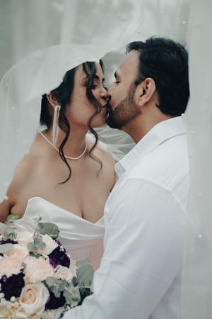 Bride and groom share a kiss under wedding veil holding bridal bouquet.