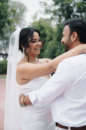 Smiling bride in white dress and veil embraces groom outdoors in park.