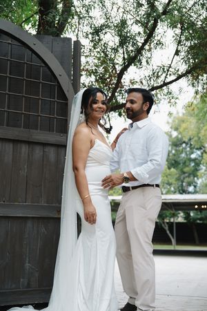 Bride in white gown and veil poses with groom near wooden arched door.