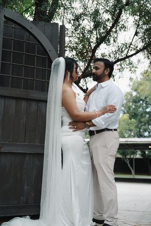 Bride and groom share romantic moment by rustic wooden gate under trees.