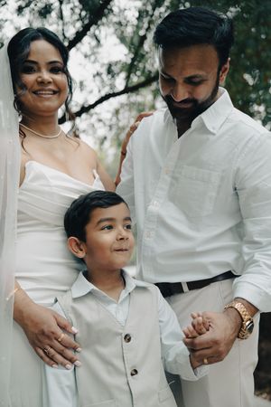 Smiling family portrait of a bride, groom, and young son standing together outdoors under trees.