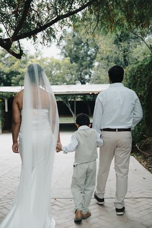 Bride in a long veil and groom walk hand-in-hand with their young son along an outdoor path.