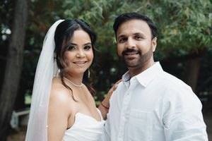 Bride in white gown and veil smiles with groom in white shirt outdoors.