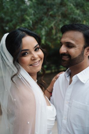 Smiling bride wearing a pearl-dotted veil stands beside her bearded groom in a white shirt outdoors.