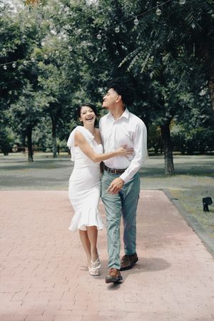 Laughing bride in white dress embraces her groom while walking along a brick path in a park.