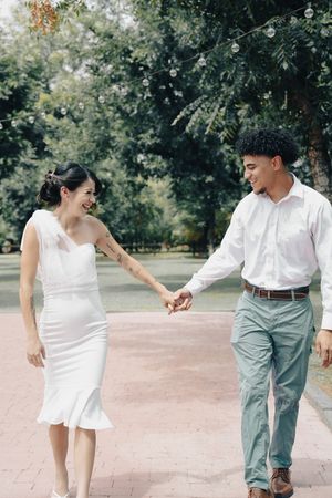 Interracial couple holding hands and laughing while walking on a brick path in a lush green park.
