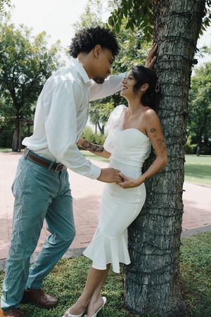 Groom leans against a tree gazing at his smiling bride in a white strapless dress outdoors.