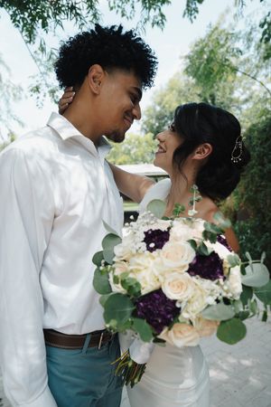 Smiling interracial couple gazes at each other lovingly, bride holding a white and purple bouquet.